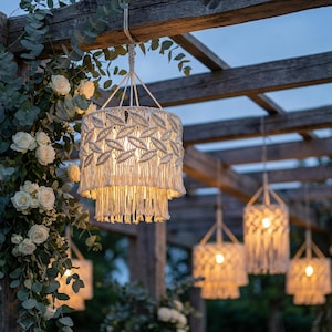 May include: Cream-colored macrame pendant lights hanging from a wooden pergola. The lights have a tiered design with a fringe at the bottom. White roses and greenery decorate the structure, creating a soft, romantic ambiance.