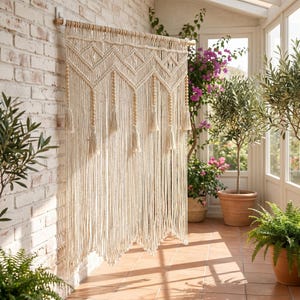 May include: Cream-colored macrame wall hanging with geometric patterns and tassels. The handmade textile art piece is suspended from a wooden dowel and hangs in front of a white brick wall. The macrame curtain adds a bohemian touch to the room.