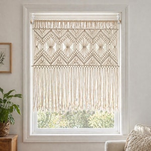 May include: A beige macrame window curtain with a geometric diamond pattern and fringe. The curtain hangs in a white window frame, adding a bohemian touch to the room. The curtain is made of woven cotton.