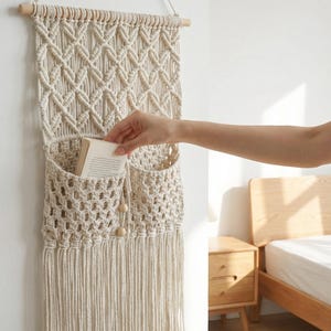 May include: Cream-colored macrame wall hanging with two pockets and a diamond pattern. The wall decor is suspended from a wooden dowel and features a fringe at the bottom. A hand is placing a book into one of the pockets.