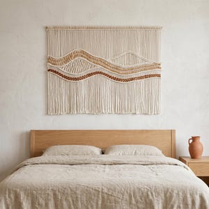 May include: A large rectangular macrame wall hanging featuring off-white cotton cord with woven mountain-like wave patterns in shades of tan, beige, and terracotta. The piece is suspended from a wooden dowel and has long fringe at the bottom.