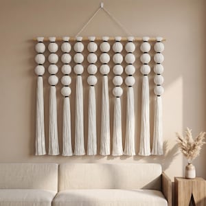 May include: A wall hanging featuring nine vertical strands of white fabric-covered spheres suspended from a horizontal wooden rod. Each strand is separated by small black accents and terminates in a long, straight white tassel. A white cord is attached to the rod for mounting.