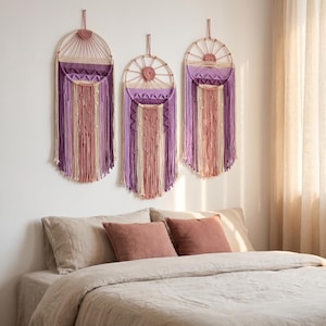 Puede incluir: Tres tapices de pared con marcos circulares de madera y diseños de macramé en tonos morado, crema y rosa empolvado. Estas piezas de arte textil incluyen patrones de nudos, flecos y pequeñas cuentas de madera que cuelgan verticalmente.