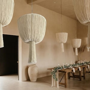 May include: Cream-coloured, fringed pendant lights hang from the ceiling in a spacious room. The lights are suspended above a long wooden table set for a gathering. A large, neutral-toned vase sits on the floor.