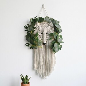 May include: A macrame wolf head wall hanging with a cream-colored design, black eyes, and a black nose. The wolf is surrounded by green leafy branches. The wall hanging is suspended from a white cord.