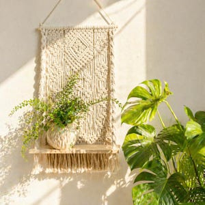 May include: A macrame wall hanging with a wooden shelf, holding a small ceramic vase with green plants. The macrame is beige with a geometric pattern. The shelf has a fringe. A large green plant is in the background.