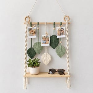 May include: A macrame photo display shelf with a wooden shelf and a hanging design. The display features leaf-shaped macrame accents in shades of green and cream, holding photos. A small potted plant and sunglasses are on the shelf.