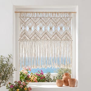 May include: A macrame window curtain in a natural beige color, featuring a geometric diamond pattern and long fringe. The curtain hangs in a white window frame, with a view of the ocean and colorful flowers.