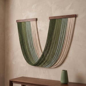 May include: A wall hanging featuring two dark brown wooden bars from which long, vertical strands of fiber drape in a deep U-shape. The strands transition in color from dark olive green to sage green and cream, creating a layered, flowing effect.