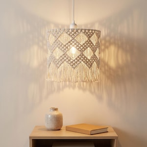 May include: A cylindrical pendant light shade crafted from off-white macrame cord featuring a repeating diamond pattern and a fringed bottom edge. The shade is suspended by a white cord and illuminates the space with a warm, soft glow.
