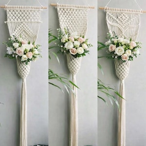 May include: Cream-colored macrame wall hanging with a wooden dowel. The macrame features a woven top section, a diamond-shaped design, and a hanging basket filled with white and pink roses and greenery. A long tassel hangs below.