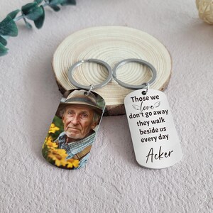 May include: A silver keychain with two tags. One tag has a photo of an older man wearing a hat and the other tag has the text "Those we love don't go away they walk beside us every day Acker".