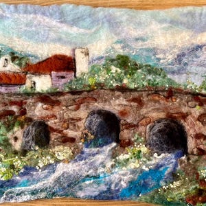 May include: A needle felted artwork depicting a stone bridge over a rushing stream. The bridge is made of brown and gray stones and has three arches. A small white building with a red roof is visible in the background. The sky is a light blue with white clouds.