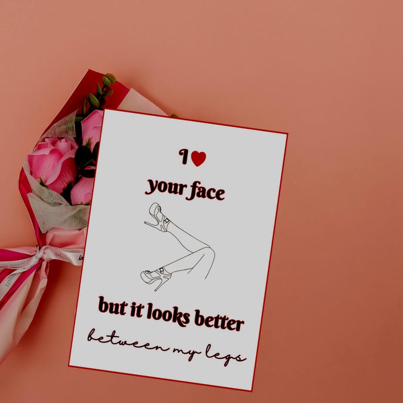 以下が含まれることがあります： 「I love your face but it looks better between my legs」のテキストとハイヒールを履いた脚のイラストが描かれたグリーティングカード。ピンクのバラの花束が背景に見えます。カードは、桃色の表面にあります。