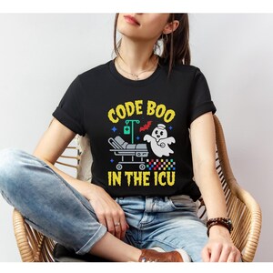May include: Black t-shirt featuring the phrase "CODE BOO IN THE ICU" in yellow and white. The design includes a ghost wearing a stethoscope, a hospital bed, a bat, and medical-themed graphics.