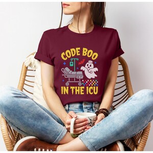 May include: A maroon t-shirt featuring the text "CODE BOO IN THE ICU" in yellow. The design includes a ghost in a nurse's hat, a hospital bed, and a bat. The shirt is made of a soft fabric.