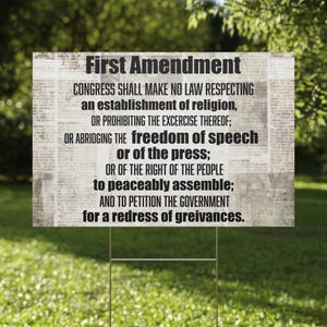 May include: A rectangular sign with the text of the First Amendment of the U.S. Constitution. The sign is white with black text and is mounted on a metal stake, displayed outdoors in a grassy area.