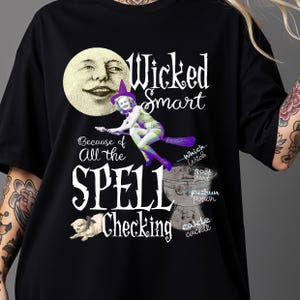 May include: Black t-shirt with a whimsical Halloween design. It features a smiling moon, a witch on a broom, and the words "Wicked Smart" and "Spell Checking." A cat and cauldron are also included in the design.