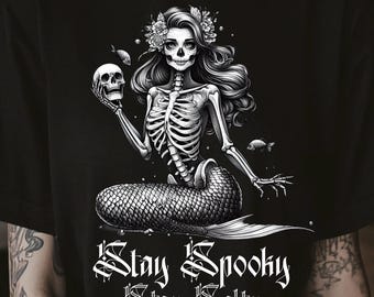 May include: Black t-shirt featuring a skeletal mermaid holding a skull, adorned with floral headwear. The design includes the phrases "Stay Spooky, Stay Salty" in a gothic font. The artwork is detailed and monochromatic, creating a spooky aesthetic.