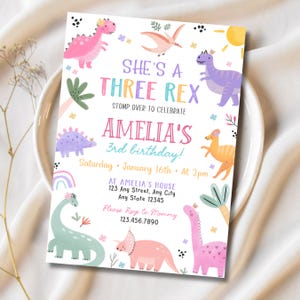 May include: A colorful birthday invitation with dinosaur illustrations. The text reads "She's a Three Rex" and "Amelia's 3rd birthday!" with details for the party. The design includes a rainbow, sun, and various dinosaur characters in pink, purple, and green.