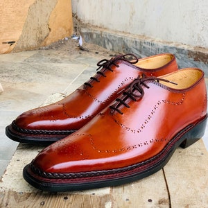 May include: A pair of brown leather dress shoes with brogue detailing. The shoes have a glossy finish, dark brown laces, and a decorative stitched border around the sole. The soles are black with a red accent.