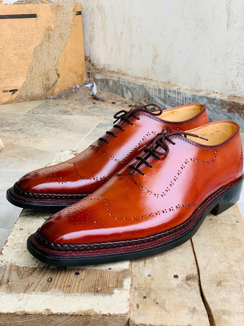 May include: A pair of brown leather dress shoes with brogue detailing. The shoes have a glossy finish, dark brown laces, and a decorative stitched border around the sole. The soles are black with a red accent.