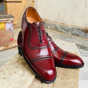 May include: A pair of burgundy leather dress shoes with dark brown accents and laces. The shoes feature a classic Oxford design with decorative perforations and a polished finish. The shoes are displayed on a wooden surface.
