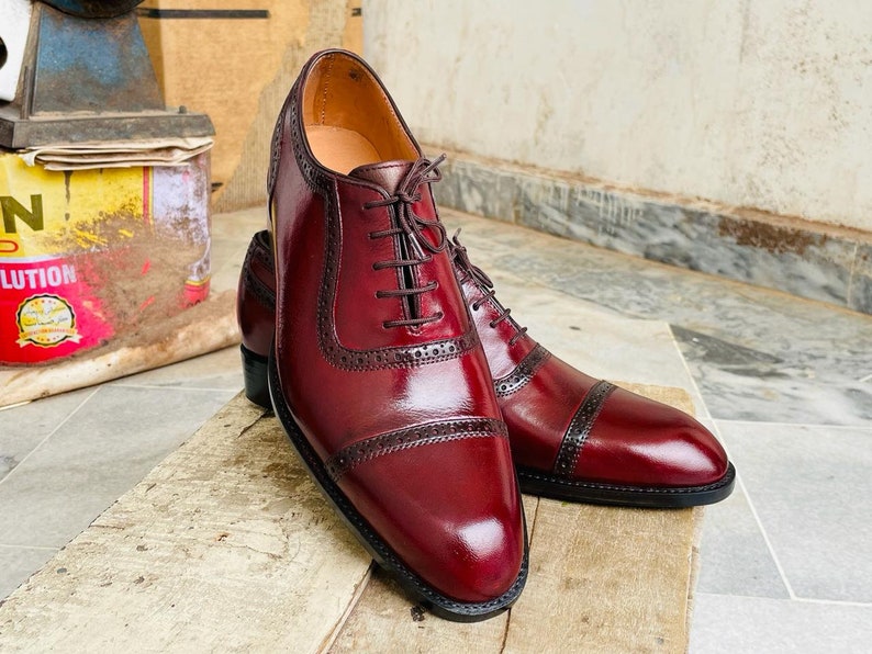 May include: A pair of burgundy leather dress shoes with dark brown accents and laces. The shoes feature a classic Oxford design with decorative perforations and a polished finish. The shoes are displayed on a wooden surface.
