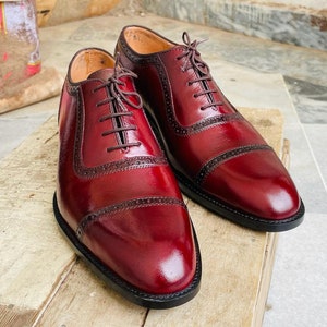 May include: A pair of burgundy leather dress shoes with a glossy finish. The shoes feature a classic Oxford design with decorative perforations and dark brown laces. The shoes are displayed on a wooden surface.