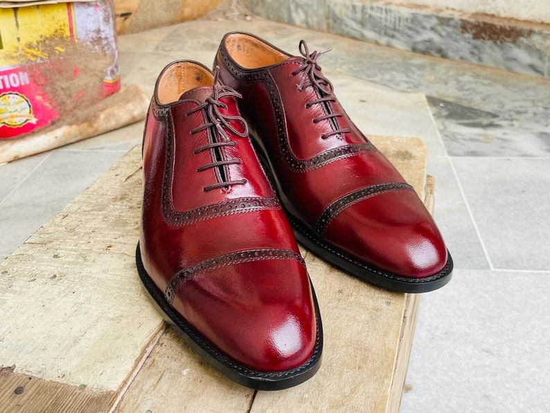 May include: A pair of burgundy leather dress shoes with a glossy finish. The shoes feature a classic Oxford design with decorative perforations and dark brown laces. The shoes are displayed on a wooden surface.