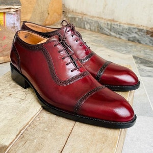 May include: A pair of burgundy leather dress shoes with cap toes and decorative perforations. The shoes have dark brown laces and a black sole. The shoes are displayed on a wooden surface.