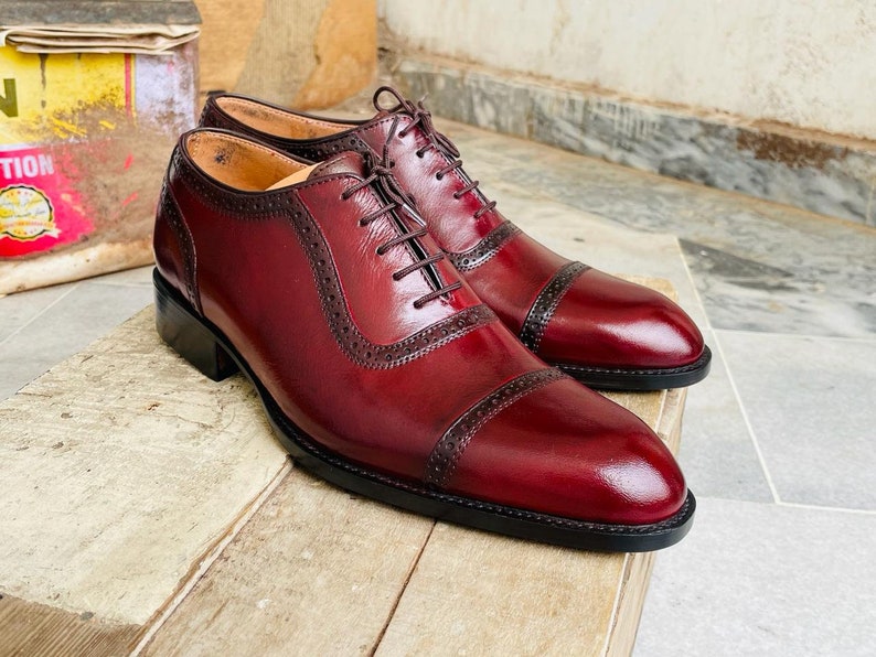 May include: A pair of burgundy leather dress shoes with cap toes and decorative perforations. The shoes have dark brown laces and a black sole. The shoes are displayed on a wooden surface.