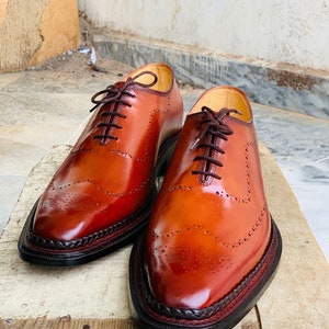 May include: Two polished, brown leather dress shoes with brogue detailing. The shoes have dark brown laces and a braided trim around the soles. The leather has a glossy finish. The shoes are displayed on a wooden surface.