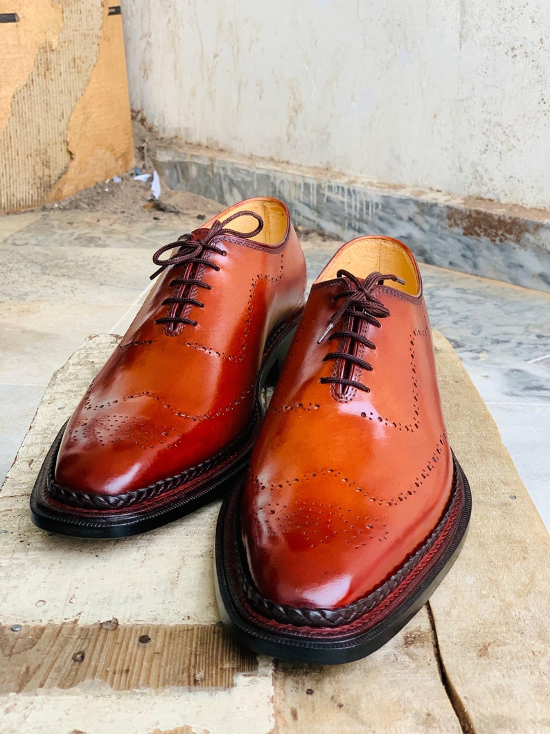 May include: Two polished, brown leather dress shoes with brogue detailing. The shoes have dark brown laces and a braided trim around the soles. The leather has a glossy finish. The shoes are displayed on a wooden surface.