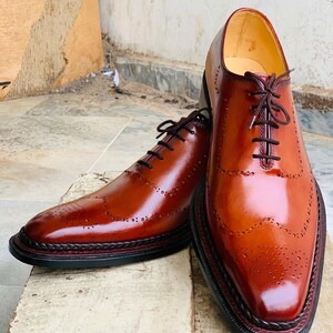 May include: Smart brown leather brogue shoes with dark brown laces. The shoes have a polished finish and decorative perforations. They feature a stitched welt and a black sole with a dark red trim. The shoes are displayed on a wooden surface.