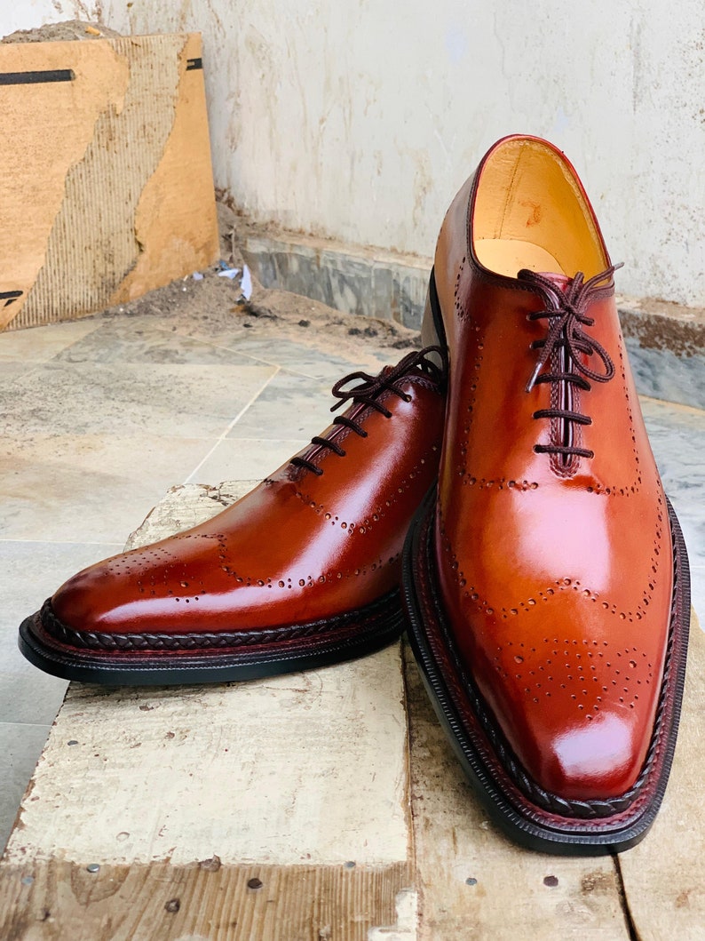 May include: Smart brown leather brogue shoes with dark brown laces. The shoes have a polished finish and decorative perforations. They feature a stitched welt and a black sole with a dark red trim. The shoes are displayed on a wooden surface.