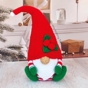 May include: A knitted gnome figurine featuring a tall red hat with a white pom-pom, a white beard, and a tan nose. The gnome wears green mittens and green boots, with red and white striped sleeves and legs. It has a holly leaf and berry accent.