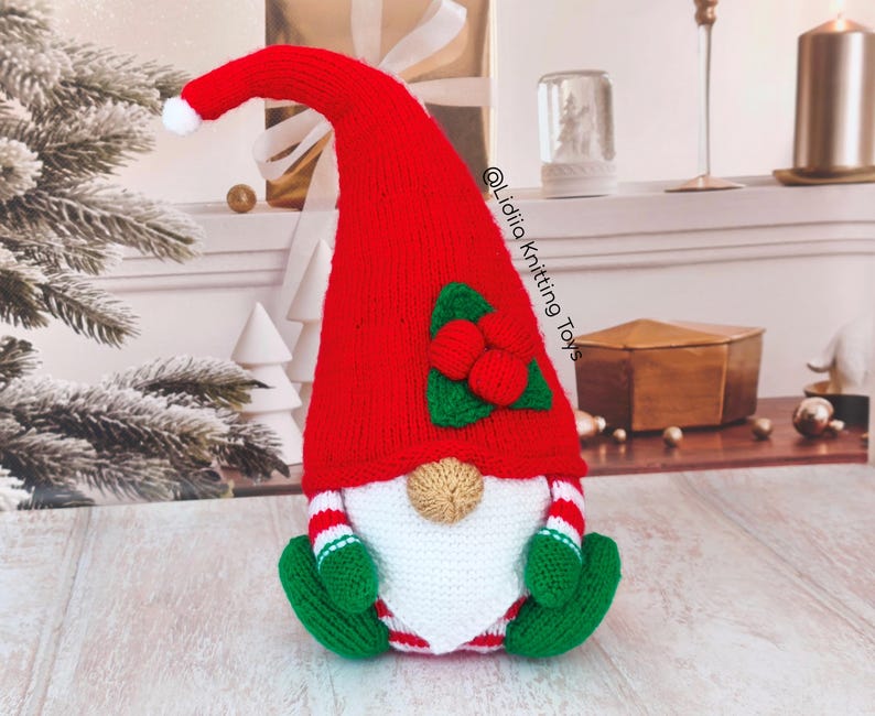 May include: A knitted gnome figurine featuring a tall red hat with a white pom-pom, a white beard, and a tan nose. The gnome wears green mittens and green boots, with red and white striped sleeves and legs. It has a holly leaf and berry accent.