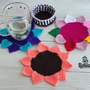 Flat lay of three vibrant flower-shaped knitted coasters and a striped brown and white storage basket on a white wooden background.