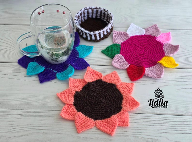 Flat lay of three vibrant flower-shaped knitted coasters and a striped brown and white storage basket on a white wooden background.