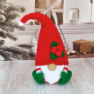 May include: A knitted gnome figurine featuring a tall, pointed red hat with a white pom-pom, adorned with green leaves and red berries. The gnome has a white beard, a tan nose, green mittens, and red and white striped sleeves. Text reads @Lidiia Knitting Toys.