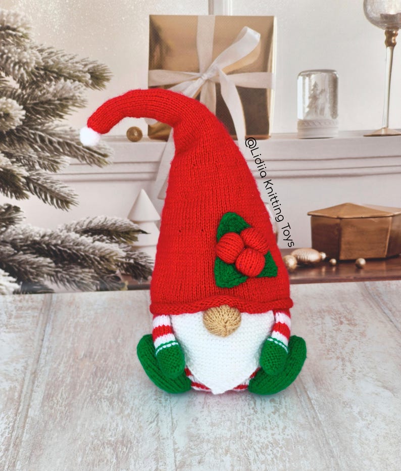 May include: A knitted gnome figurine featuring a tall, pointed red hat with a white pom-pom, adorned with green leaves and red berries. The gnome has a white beard, a tan nose, green mittens, and red and white striped sleeves. Text reads @Lidiia Knitting Toys.