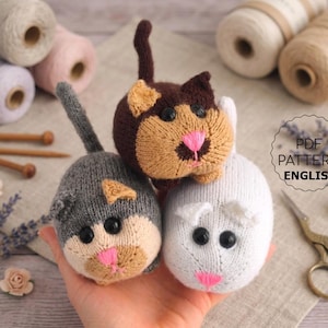 May include: Three small, round, hand-knitted cat toys in grey, dark brown, and white. Each cat features black plastic eyes, a stitched pink nose, and a small tail. The image includes text that reads PDF PATTERN ENGLISH.