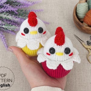 May include: Two handmade knitted chicken toys. The chickens have white bodies, red combs, black eyes, and yellow beaks. One chicken has a yellow base, and the other has a red base. The image also includes a PDF pattern in English.