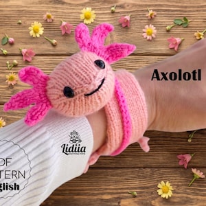 Könnte beinhalten: Ein hellrosa gestricktes Axolotl-Armband mit leuchtend rosa Kiemen und schwarzen Perlenaugen. Der Artikel enthält den Text Axolotl, Lidiia Knitting Toys und PDF PATTERN English. Das Design umfasst ein breites gestricktes Band für das Handgelenk.