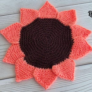 May include: A round, hand-knitted sunflower-shaped coaster or mat featuring a dark brown circular center surrounded by twelve pointed, coral-pink petals. The text Lidiia KNITTING TOYS is printed in the upper right corner of the image.