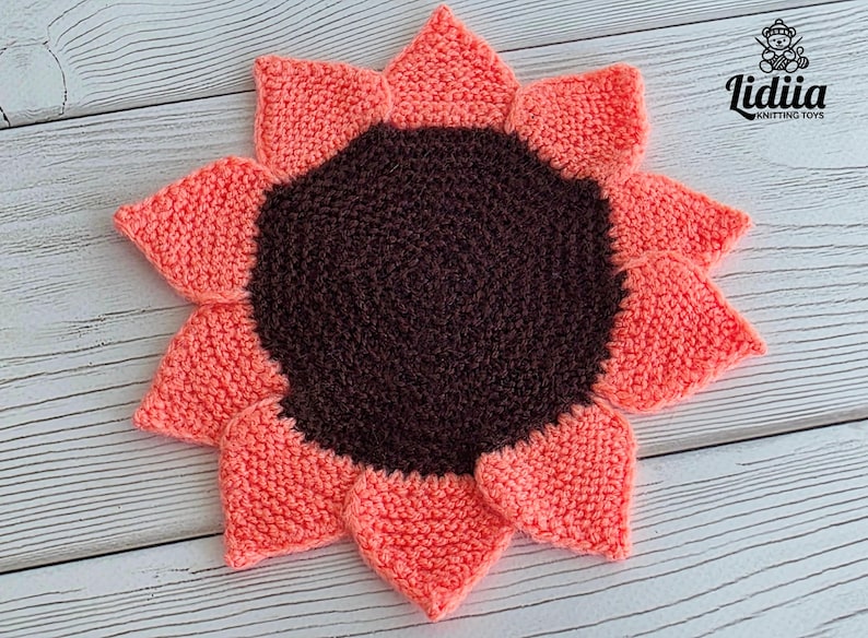 May include: A round, hand-knitted sunflower-shaped coaster or mat featuring a dark brown circular center surrounded by twelve pointed, coral-pink petals. The text Lidiia KNITTING TOYS is printed in the upper right corner of the image.