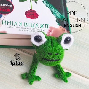 May include: A green knitted frog bookmark with white-rimmed eyes and long legs. The bookmark rests on a book with English and Ukrainian text. The text "PDF PATTERN ENGLISH" is also visible, indicating a pattern.