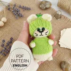 Puede incluir: Un juguete de ratón tejido a mano, en tonos verdes y blancos, que lleva un gorro a juego con detalles de orejas. El juguete tiene ojos y bigotes negros. La imagen incluye el texto "PDF PATTERN ENGLISH".