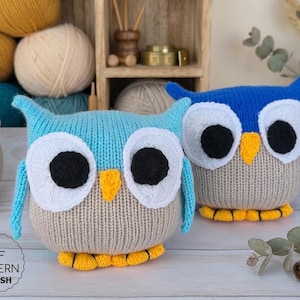 May include: Two knitted owl plush toys. One is light blue, the other is dark blue. Both have large white eyes with black pupils, yellow beaks, and yellow feet. The owls are sitting on a light-colored wooden surface.
